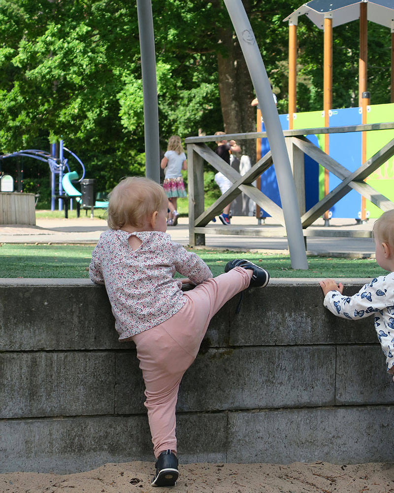 A young toddler tries hard to overstep a fence, she seems determined to climb over, towards the playground in the background.